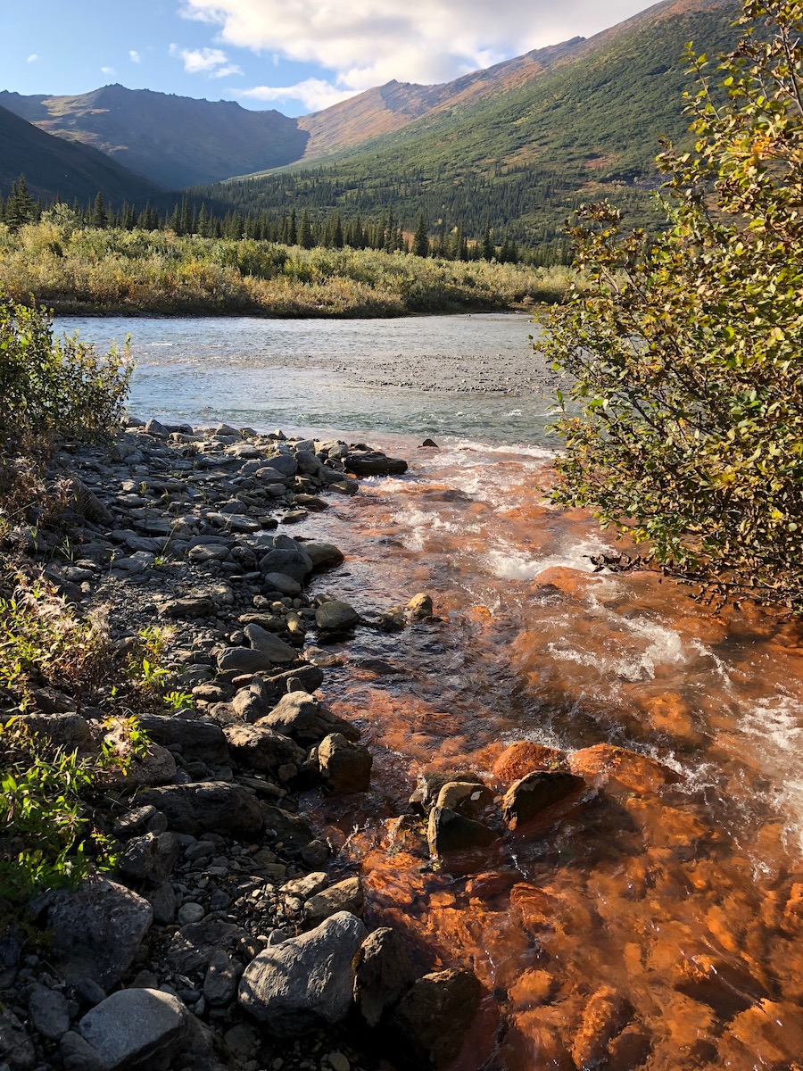A rusty stream flows into a clear river with mountains in the background.